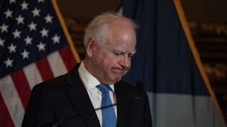 Tim Walz frowns, looking down, with an American flag in back of him.