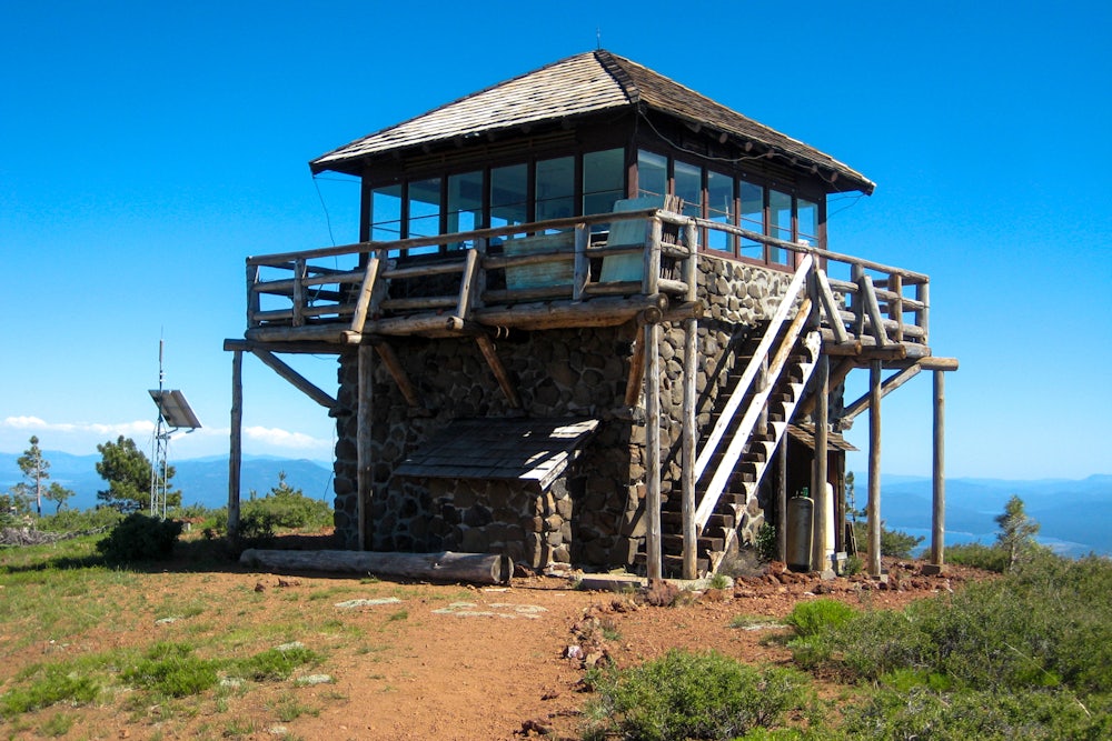 A stone and wooden tower structure sits on a sparsely vegetated bit of land overlooking mountains.