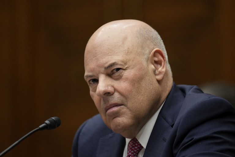 A bald man wearing a suit squints as he sits before a microphone.