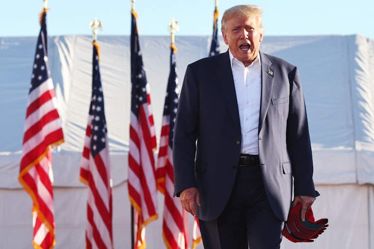 Donald Trump stands in front of five large U.S. flags, wearing a suit and holding seeral red caps in his left hand. He looks like he is yelling at the camera, or perhaps a crowd not shown in the photo.