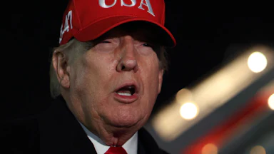 Donald Trump squints and speaks while standing next to Air Force One. He is wearing a red baseball cap that says "USA" in white letters
