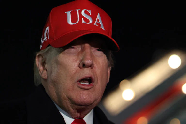 Donald Trump squints and speaks while standing next to Air Force One. He is wearing a red baseball cap that says "USA" in white letters