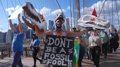 A demonstrator holds two replicas of dinosaur heads in his hands while wearing a sign reading "Don't Be a Fossil Fool!!"