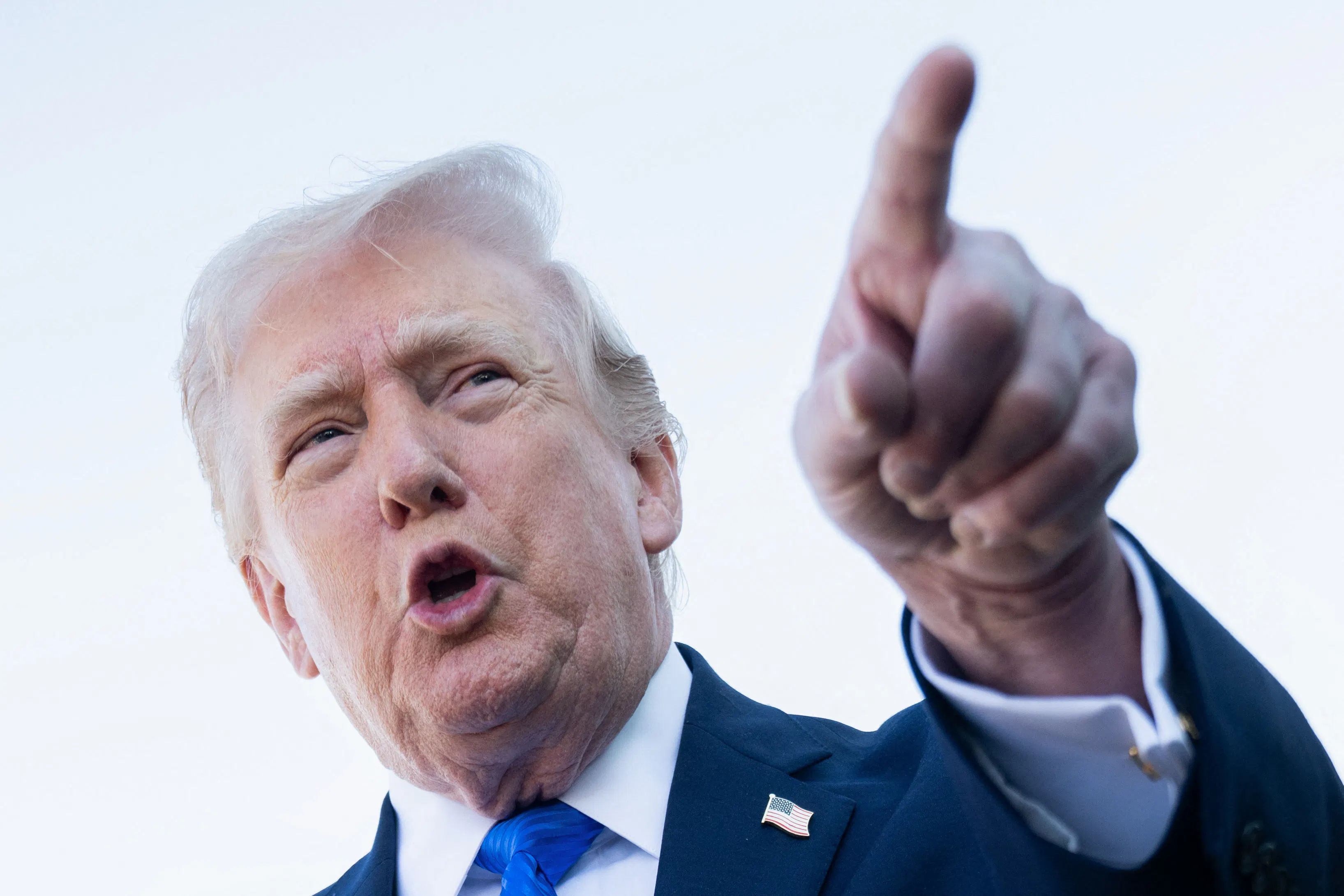 Donald Trump, wearing a blue suit and blue tie, points a finger forward with his mouth open.