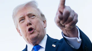 Donald Trump, wearing a blue suit and blue tie, points a finger forward with his mouth open.