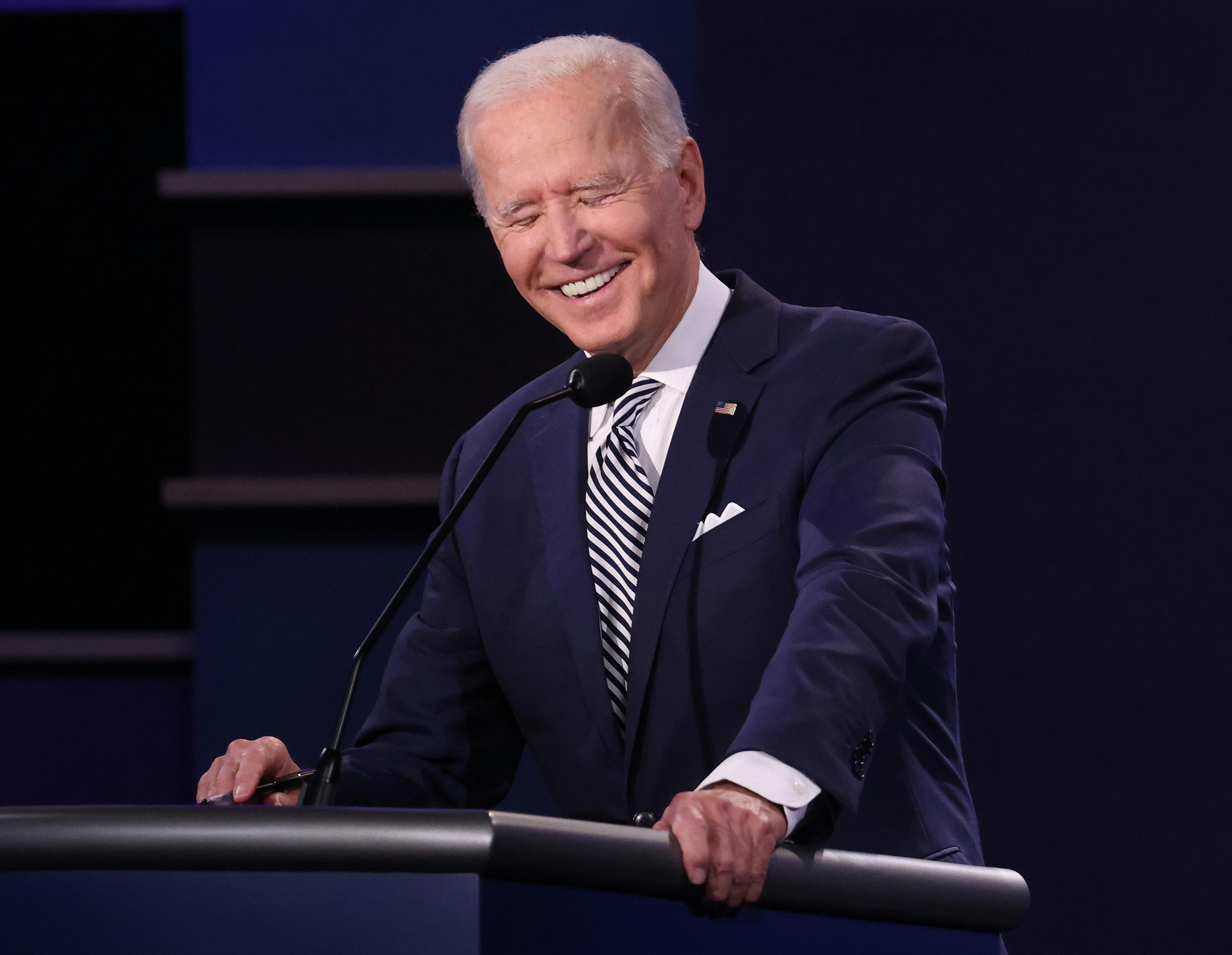 Joe Biden grasps a lectern and laughs, closing his eyes and showing pearly white teeth. 