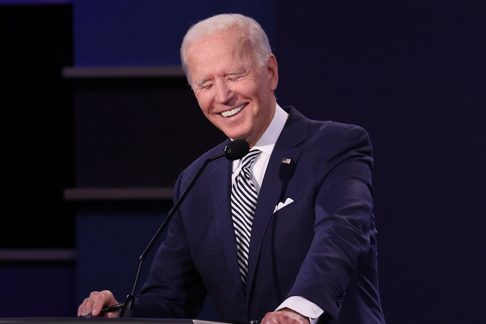 Joe Biden grasps a lectern and laughs, closing his eyes and showing pearly white teeth.