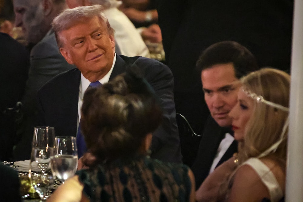 Trump sits at a table, wearing a suit, next to Marco Rubio and Jeanette Rubio.