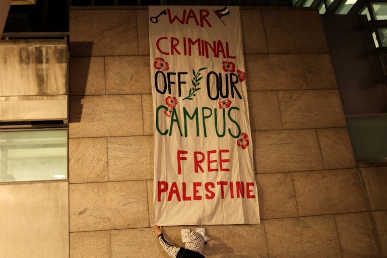 A protester wearing a keffiyeh adjusts a banner that says "WAR CRIMINAL OFF OUR CAMPUS FREE PALESTINE."