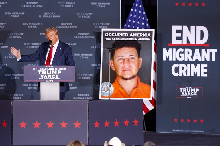 Donald Trump gestures while speaking at a podium in front of signs that say “End Migrant Crime” and “Deport Illegals Now”