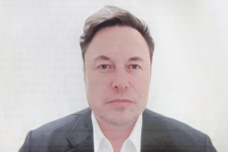 Elon Musk appears on a video screen wearing a gray suit (slightly pixelated)