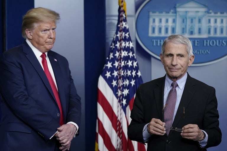 Donald Trump stands to the side, looking serious, and Dr. Anthony Fauci stands closer to the center of the White House briefing room, holding his glasses in his hands and pursing his lips.