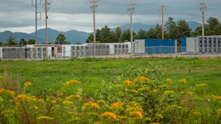 A meadow with wildflowers in the foreground with power lines and low buildings and mountains in the background.