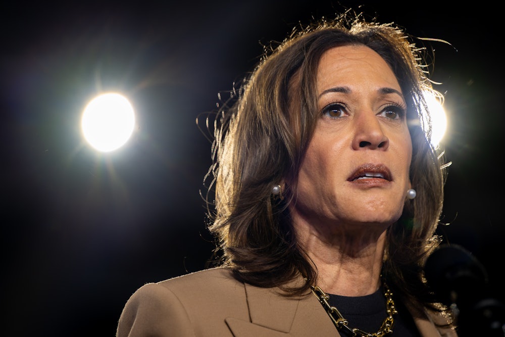 Kamala Harris, wearing a tan pantsuit, stares as stage lights shine behind her.