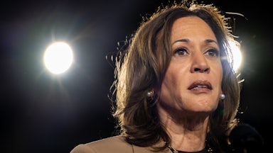Kamala Harris, wearing a tan pantsuit, stares as stage lights shine behind her.