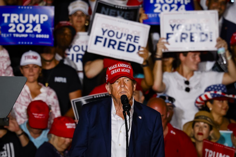 Donald Trump at a mic. People in the crowd in the stands behind him hold signs up reading "You're Fired!" and "Trump 2024."