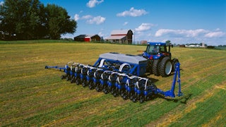 A blue tractor drives over a field with a red barn in the background.