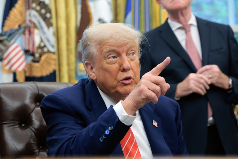 Donald Trump points at something or someone off camera, while speaking and sitting at his desk in the Oval Office of the White House.