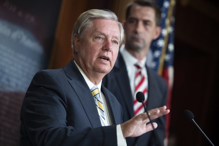 Senator Lindsey Graham wears a suit and speaks at a podium, gesturing with his hand. A blurry Tom Cotton is in the background.