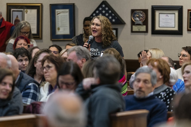 A woman holding a microphone angrily yells while others at the town hall are seated and listen.