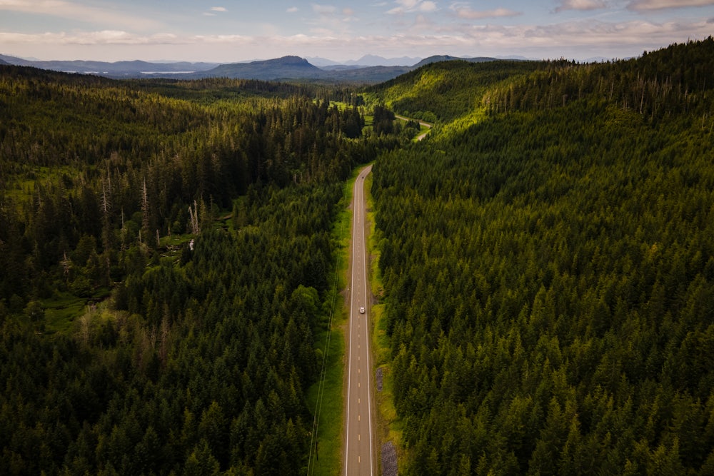 Aerial view of a truck driving through a heavily forested road with mountains in the background.