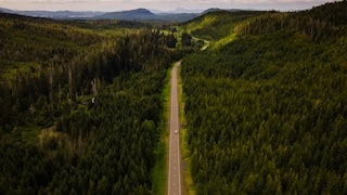 Aerial view of a truck driving through a heavily forested road with mountains in the background.