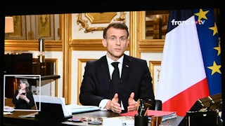 A screen displays French President Emmanuel Macron sitting at a desk and speaking