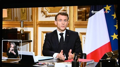 A screen displays French President Emmanuel Macron sitting at a desk and speaking