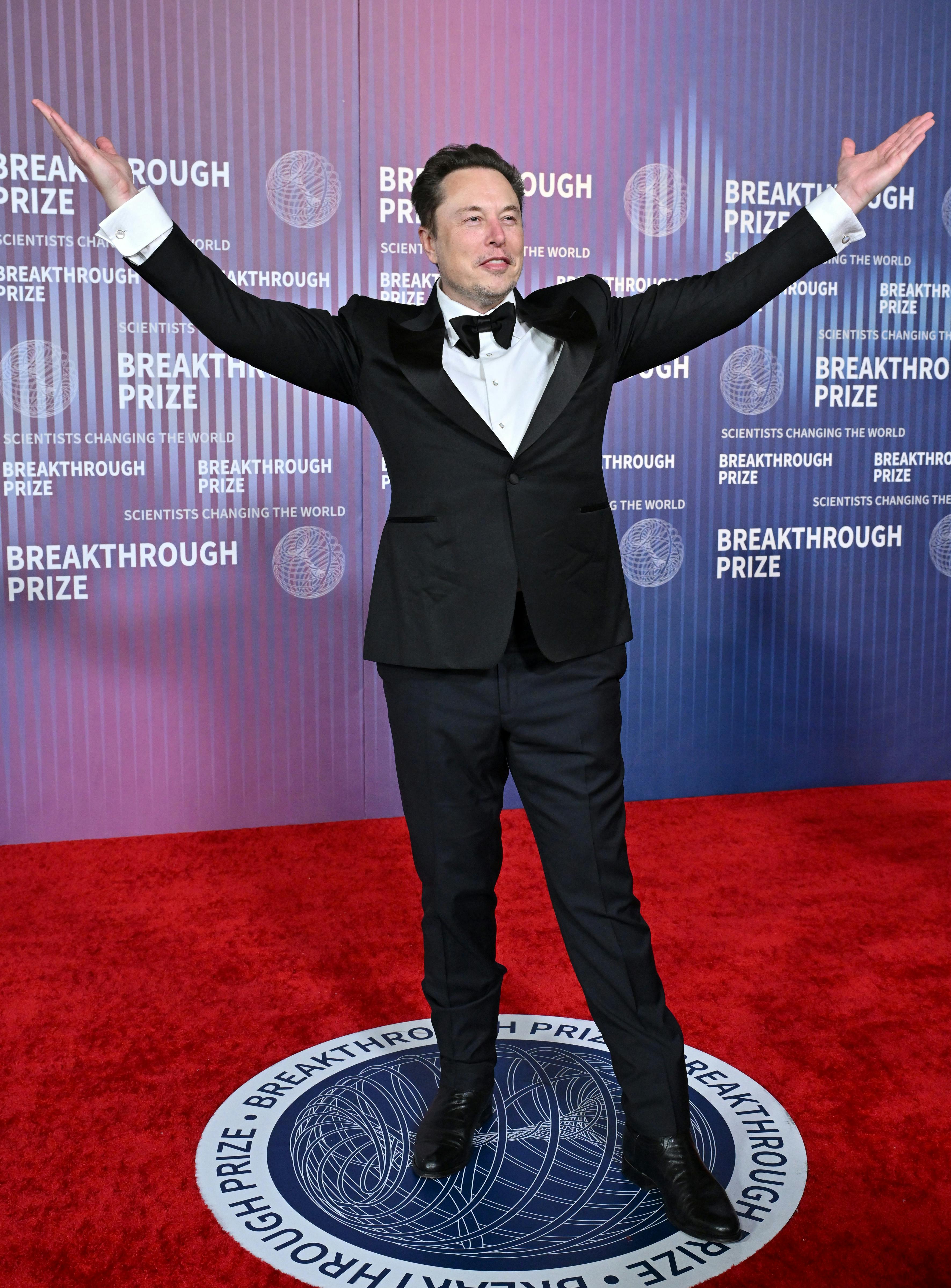 Elon Musk stands with his arms raised.