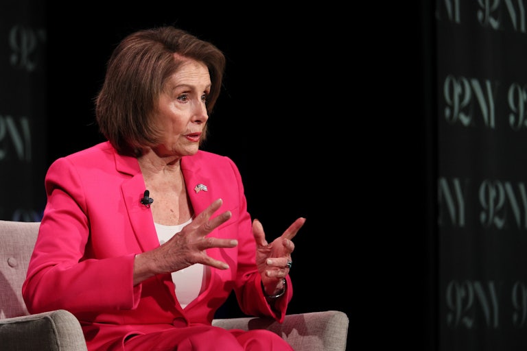 Nancy Pelosi, seated on an armchair, speaks and makes hand gestures. There is a black backdrop behind her.