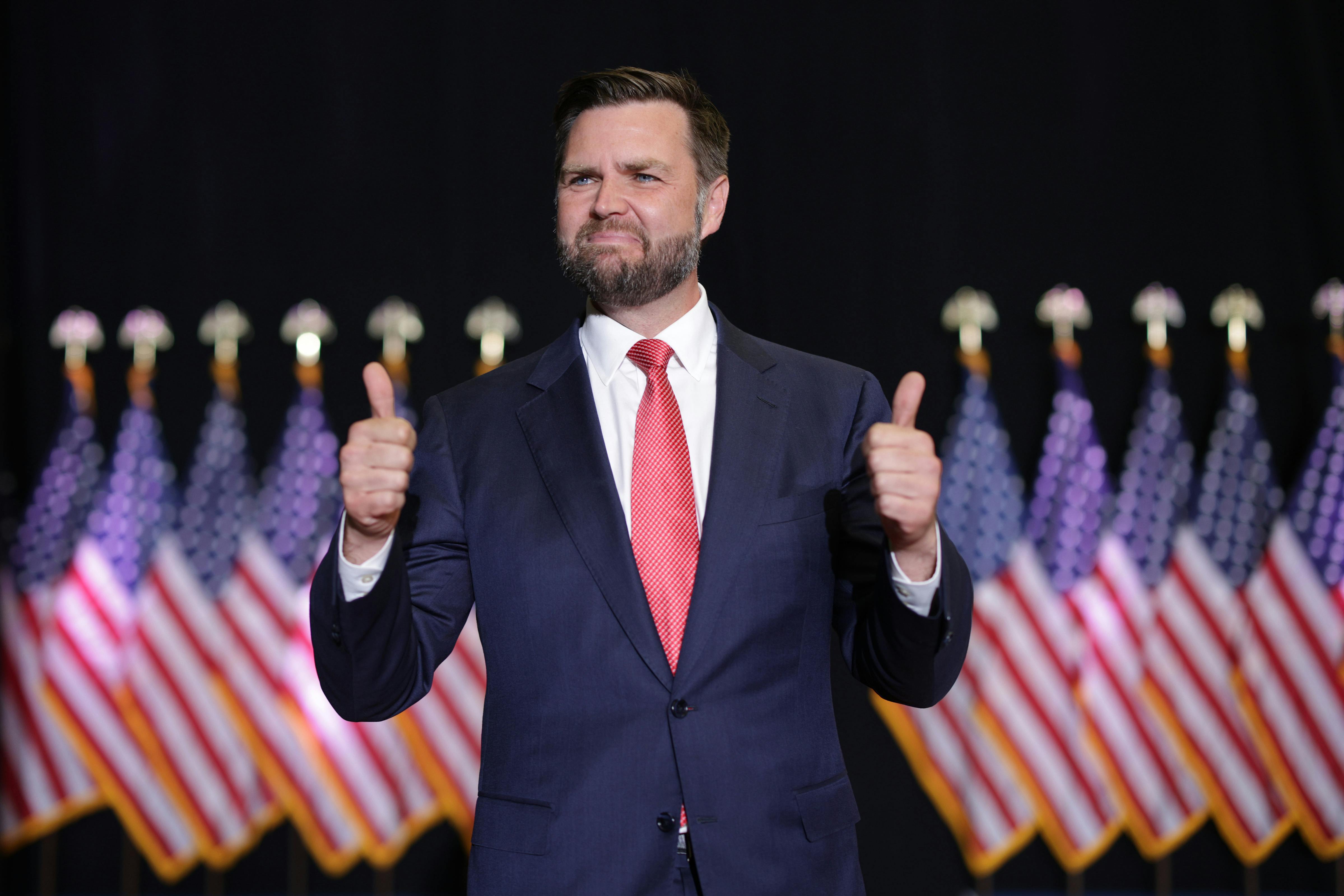 J.D. Vance awkwardly gives two thumbs ups while standing in front of a row of American flags.