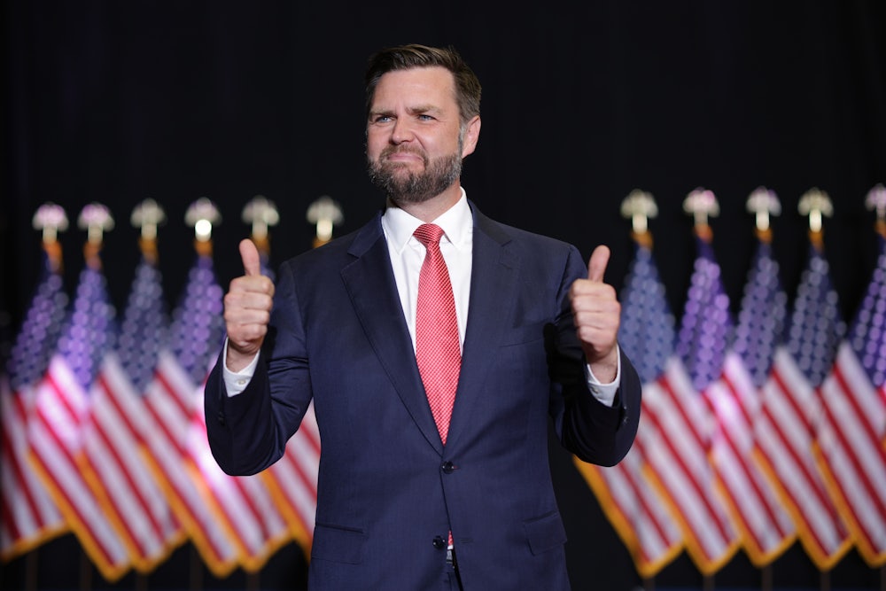 J.D. Vance awkwardly gives two thumbs ups while standing in front of a row of American flags.