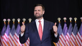 J.D. Vance awkwardly gives two thumbs ups while standing in front of a row of American flags.
