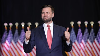 J.D. Vance awkwardly gives two thumbs ups while standing in front of a row of American flags.