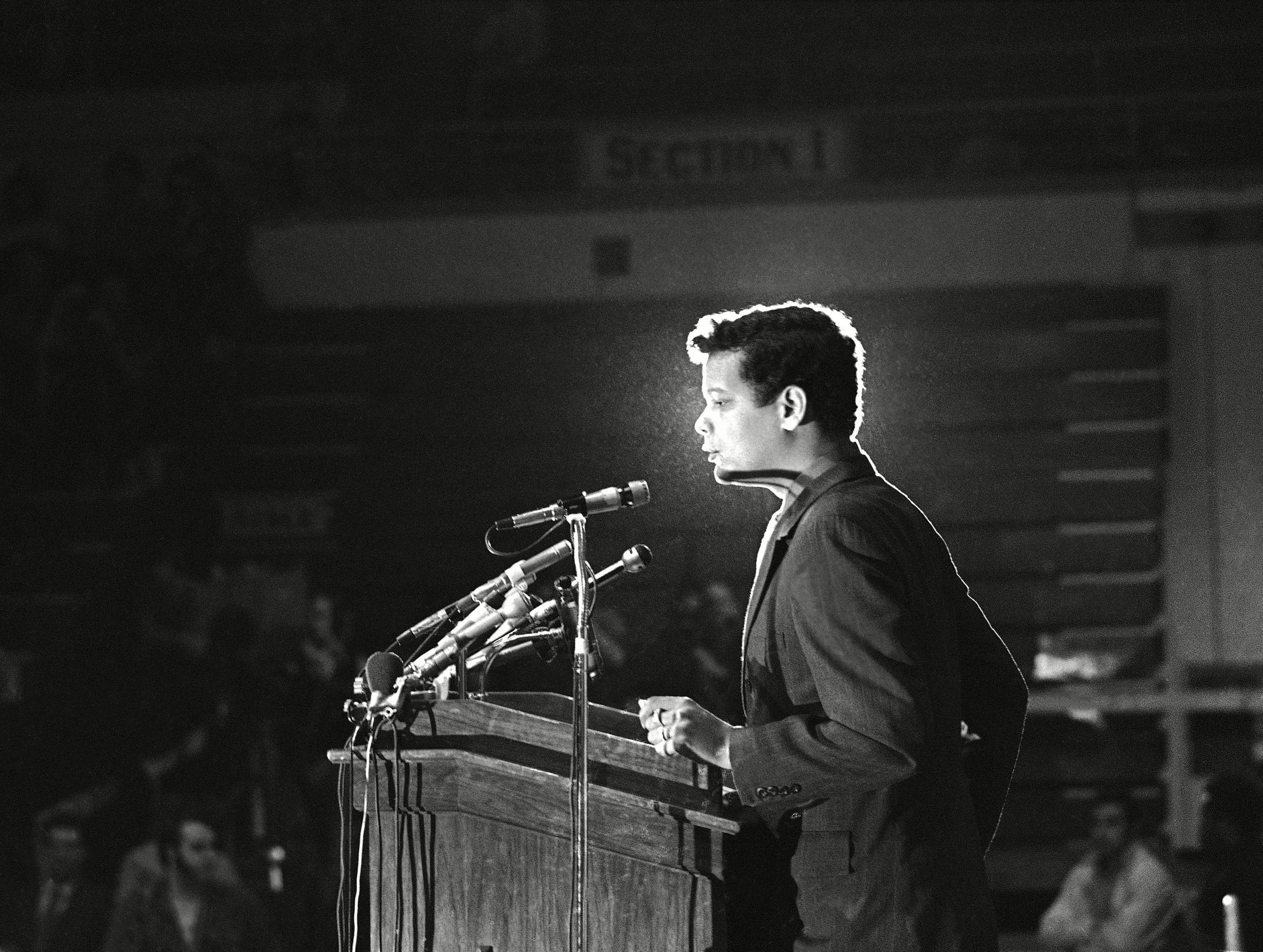 A black-and-white photograph of Julian Bond standing before a lectern equipped with microphones.