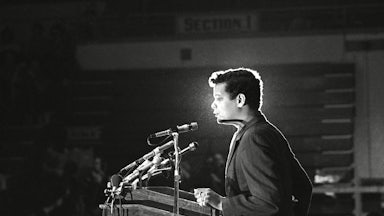 A black-and-white photograph of Julian Bond standing before a lectern equipped with microphones.