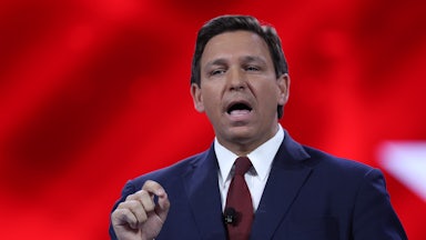 A close up of Ron DeSantis speaking to an audience in front of a red background.
