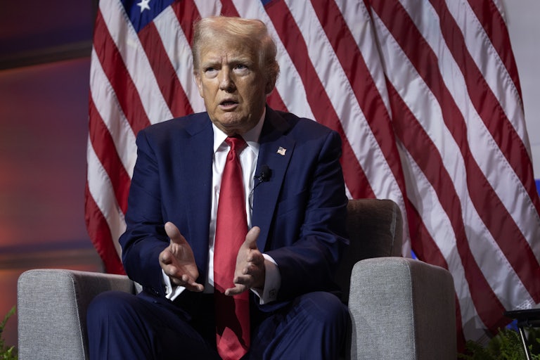 Donald Trump sits on an armchair and speaks, making hand gestures. A U.S. flag is behind him.