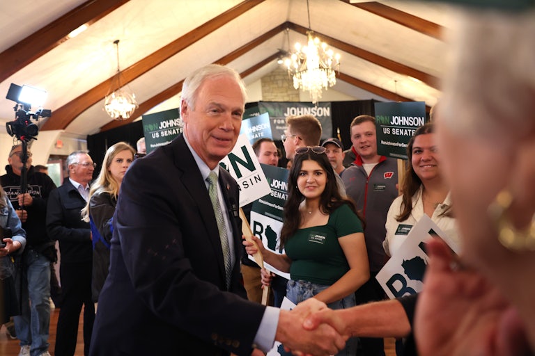 Ron Johnson shakes someone's hand as supporters stand around him with signs that read "Ron Johnson for US Senate." A camera is in the background.