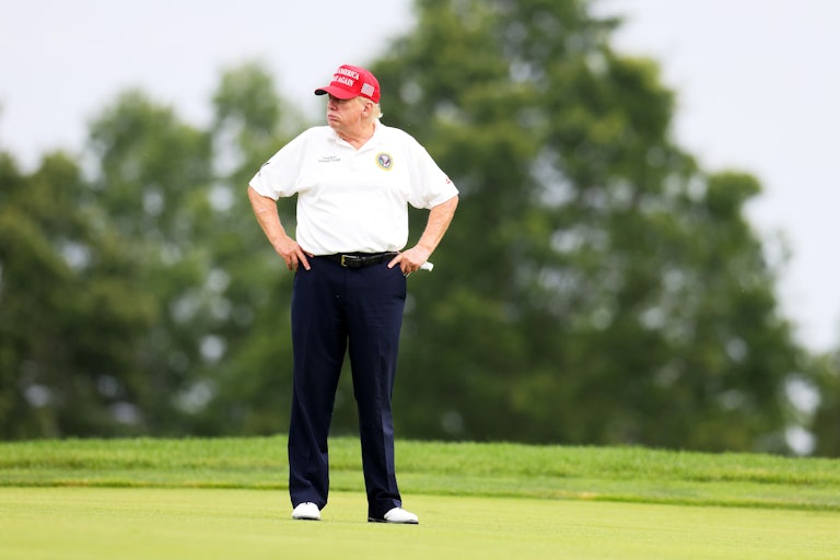 Donald Trump puts his hands on his hips while standing on the golf course. He wears a red Make America Great Again cap and looks off to the side.