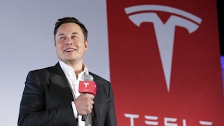 Elon Musk holds a microphone as he stands in front of a Tesla logo