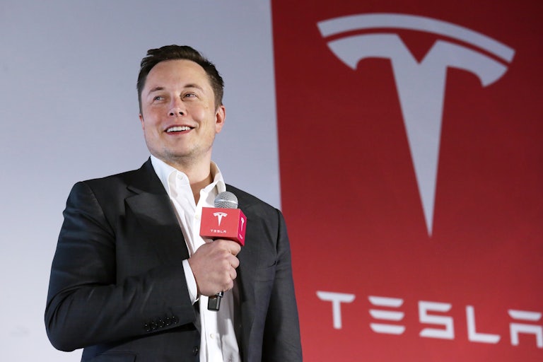 Elon Musk holds a microphone as he stands in front of a Tesla logo