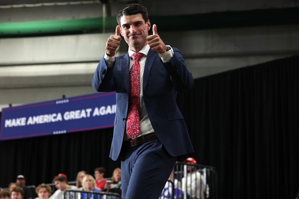 Representative Robert Bresnahan smiles on stage and gives two thumbs up to the camera. A large "Make America Great Again" banner is in the background.