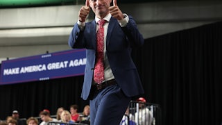 Representative Robert Bresnahan smiles on stage and gives two thumbs up to the camera. A large "Make America Great Again" banner is in the background.