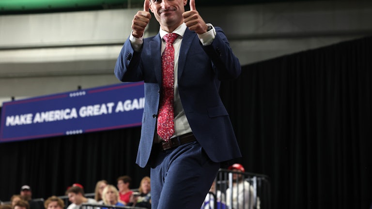 Representative Robert Bresnahan smiles on stage and gives two thumbs up to the camera. A large "Make America Great Again" banner is in the background.