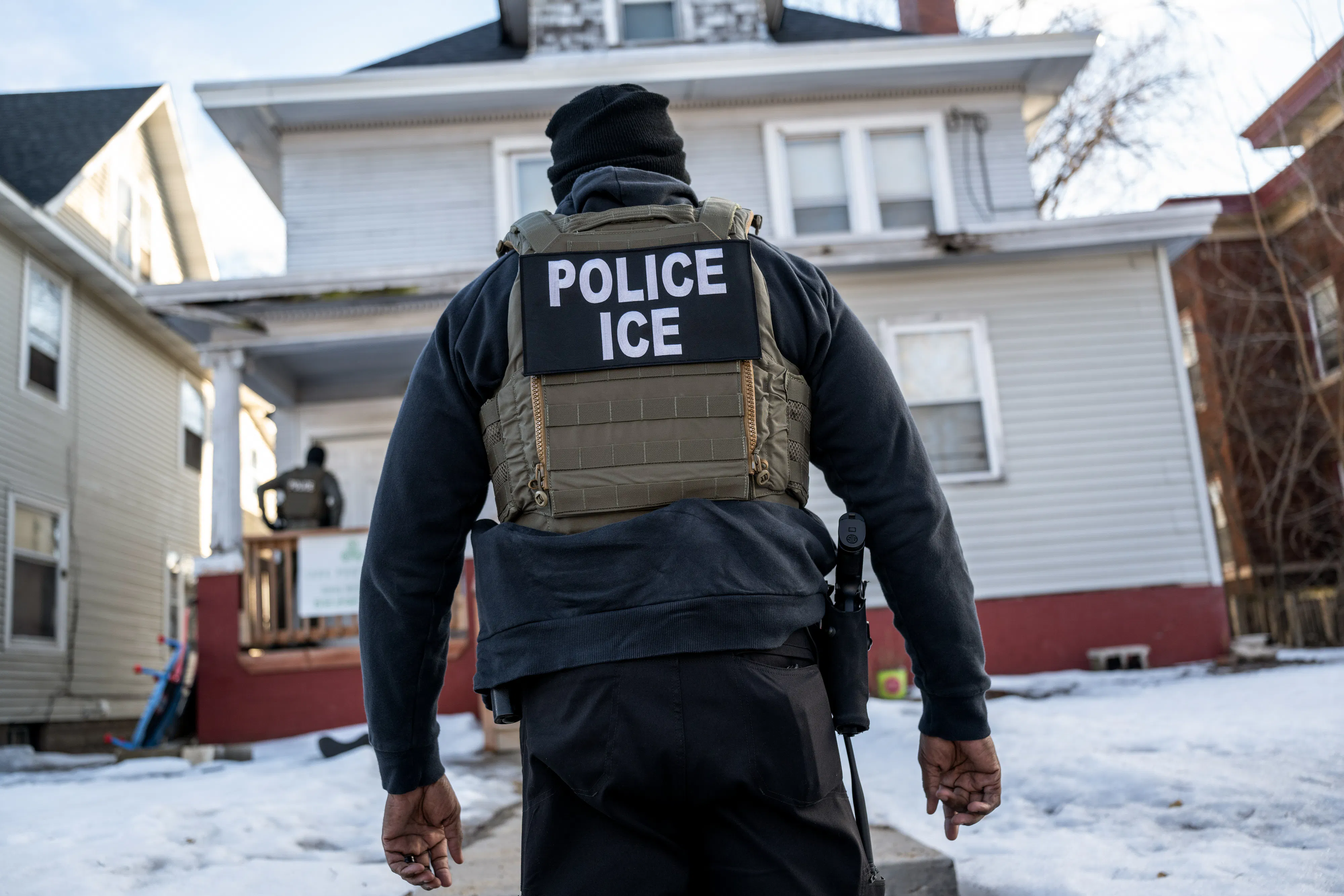 An ICE agent walks to a house in the snow (the back of his vest, with the word ICE, is pictured).