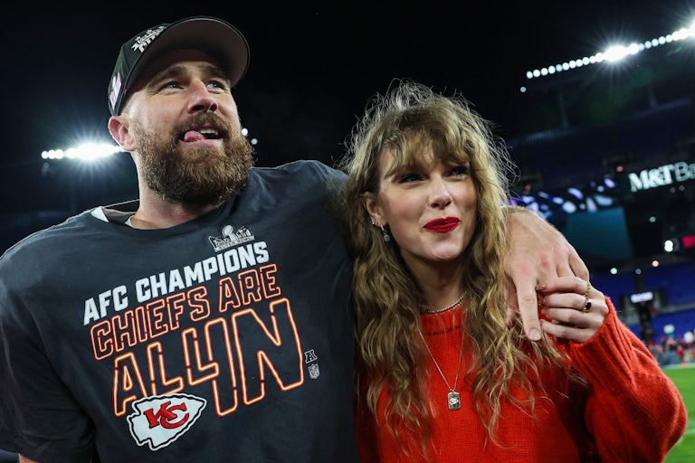 Travis Kelce throws his arm around Taylor Swift's shoulders. They're standing on the football field smiling. Kelce wears a cap and a shirt that says "AFC Champion Chiefs Are All In." Taylor Swift wears a red sweater and red lipstick.