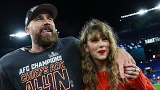 Travis Kelce throws his arm around Taylor Swift's shoulders. They're standing on the football field smiling. Kelce wears a cap and a shirt that says "AFC Champion Chiefs Are All In." Taylor Swift wears a red sweater and red lipstick.