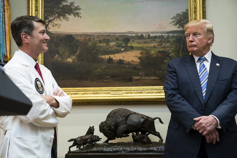 Ronny Jackson in a white doctor's coat smiles and looks over at Donald Trump who is pursing his lips staring at him