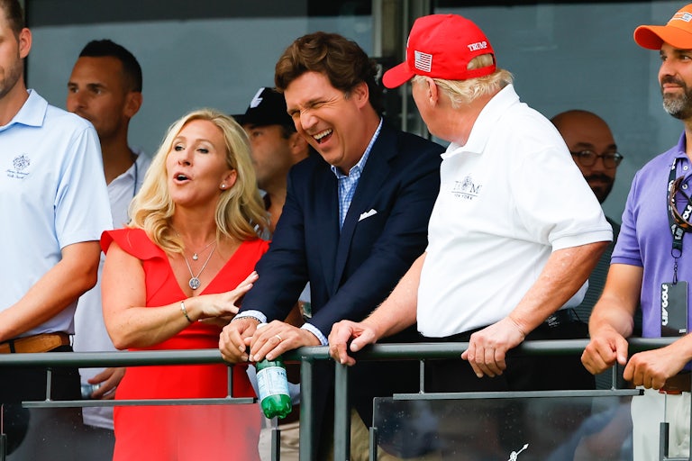 Marjorie Taylor Greene, Tucker Carlson, and Donald Trump laugh while standing on a balcony. Don Jr stands beside them.